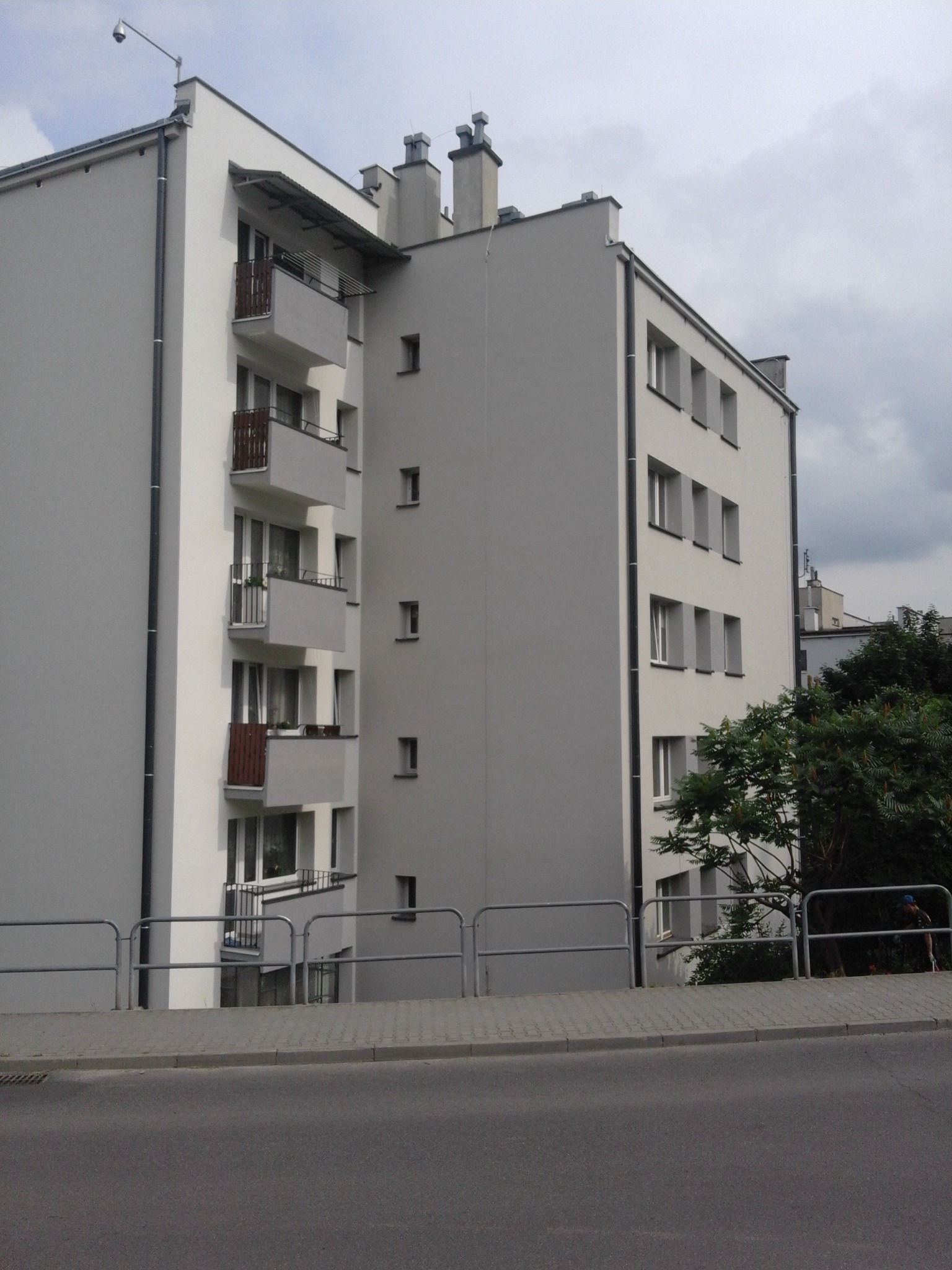 Wielopiętrowy budynek mieszkalny z balkonami po remoncie elewacji, widoczny z poziomu ulicy, z szarą elewacją i białymi ramami okiennymi.