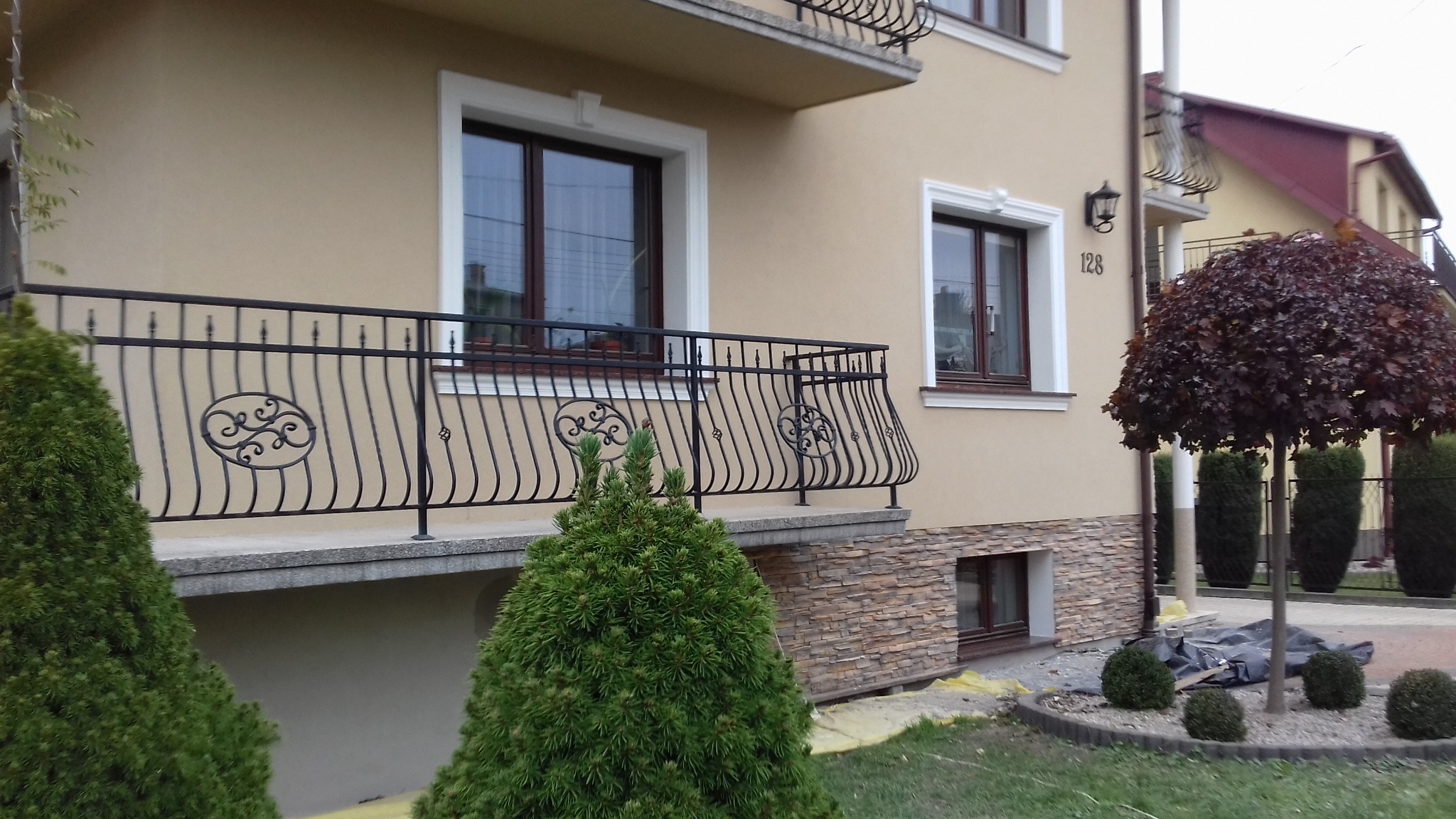 Jasna elewacja domu z balkonem o ozdobnej, metalowej balustradzie, okna z białymi ramami i numerem domu 128, kamienna podmurówka i zadbany ogród z formowanymi drzewami.