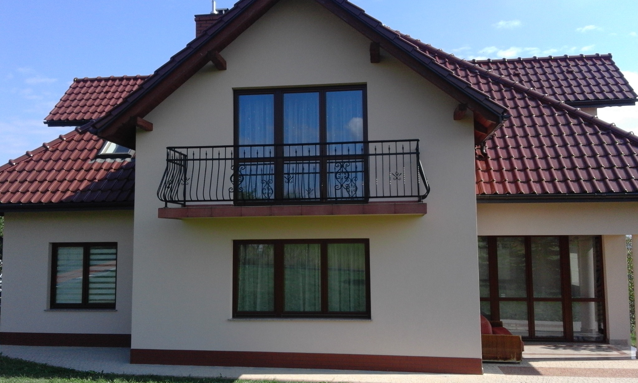 Dwupiętrowy dom z beżową elewacją, balkonem z ozdobną, kutą balustradą, brązową stolarką okienną i czerwoną dachówką