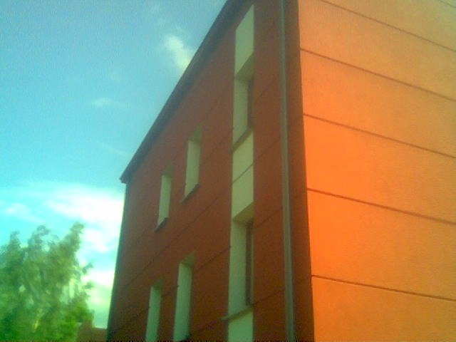 Fragment budynku z pomarańczowo-brązową elewacją, widoczne okna z białymi obramowaniami i fragment drzewa na tle błękitnego nieba.