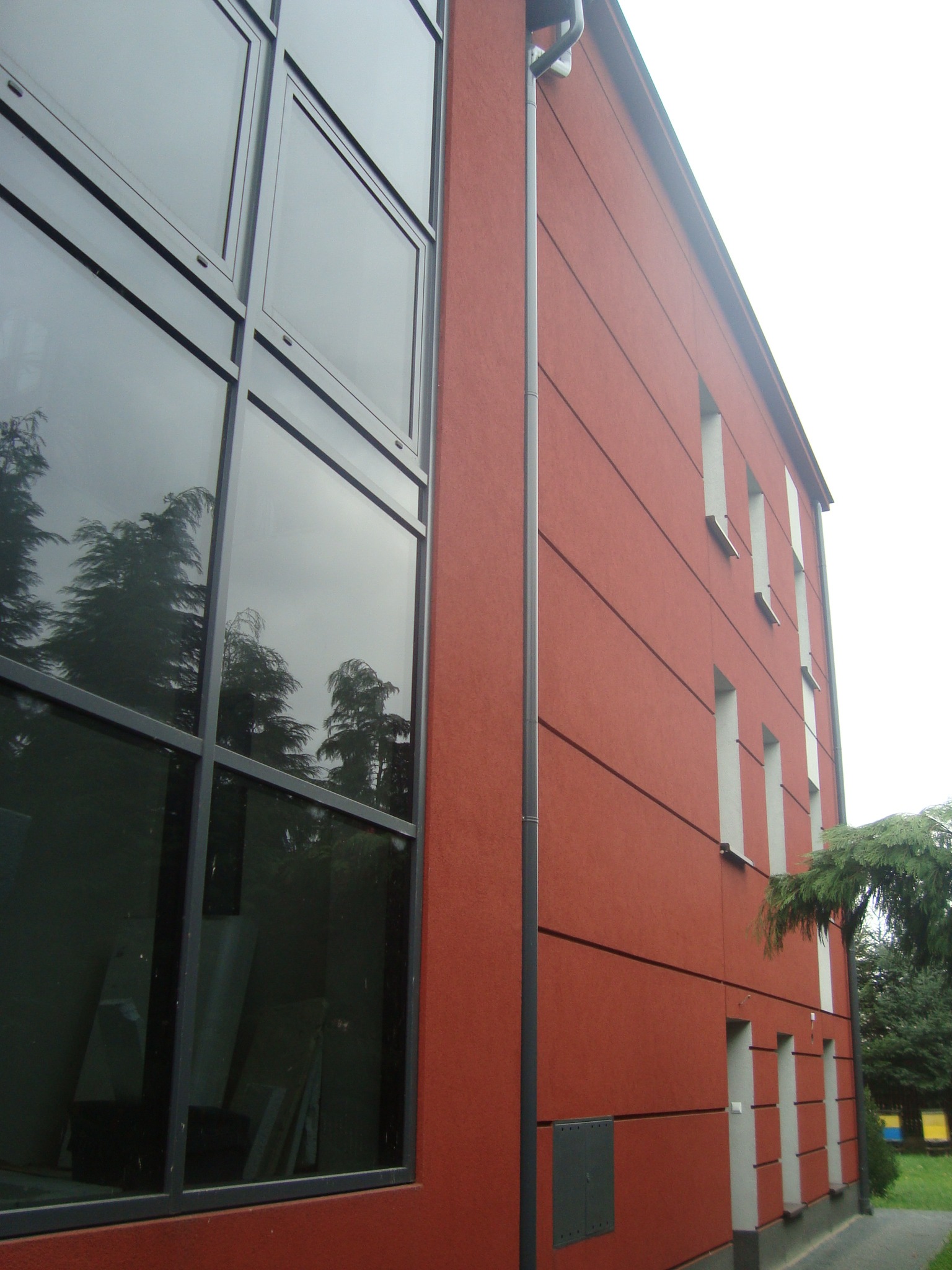 Czerwona elewacja budynku z dużymi, ciemnymi oknami, widoczne odbicie drzew w szybach, prosta architektura.