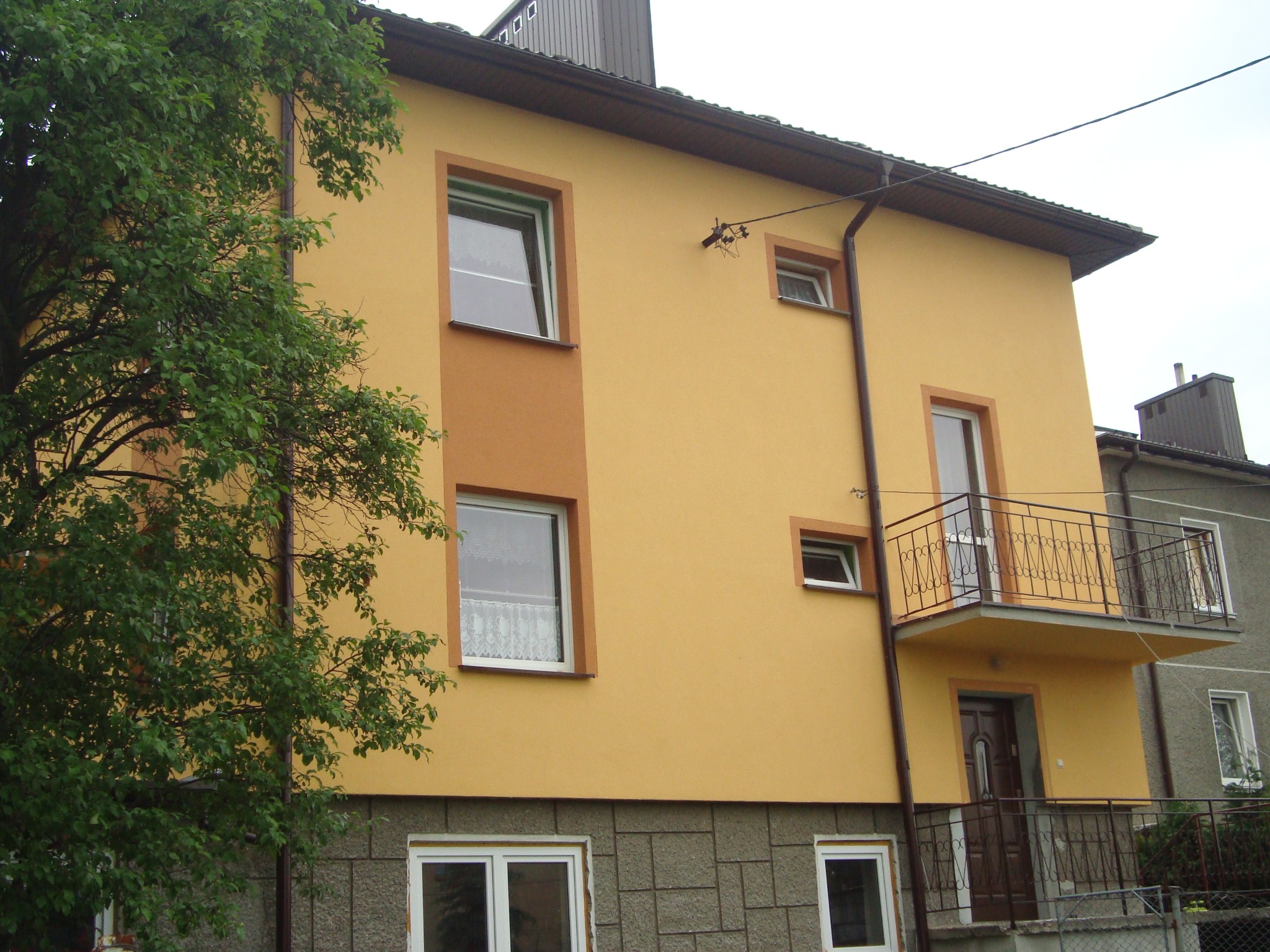 Dwupiętrowy dom z żółtą elewacją, brązowymi ramami okiennymi i balkonem z metalową balustradą, dolna część elewacji wykończona szarym kamieniem dekoracyjnym