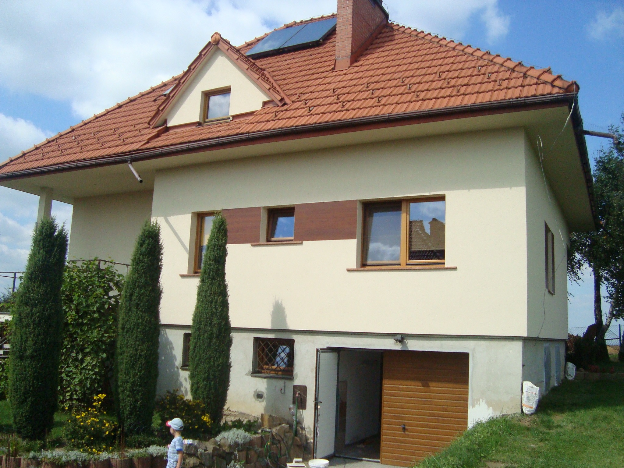 Dwukondygnacyjny dom z beżową elewacją, czerwoną dachówką i panelem słonecznym na dachu, widok z ogrodu z dzieckiem w kapeluszu.