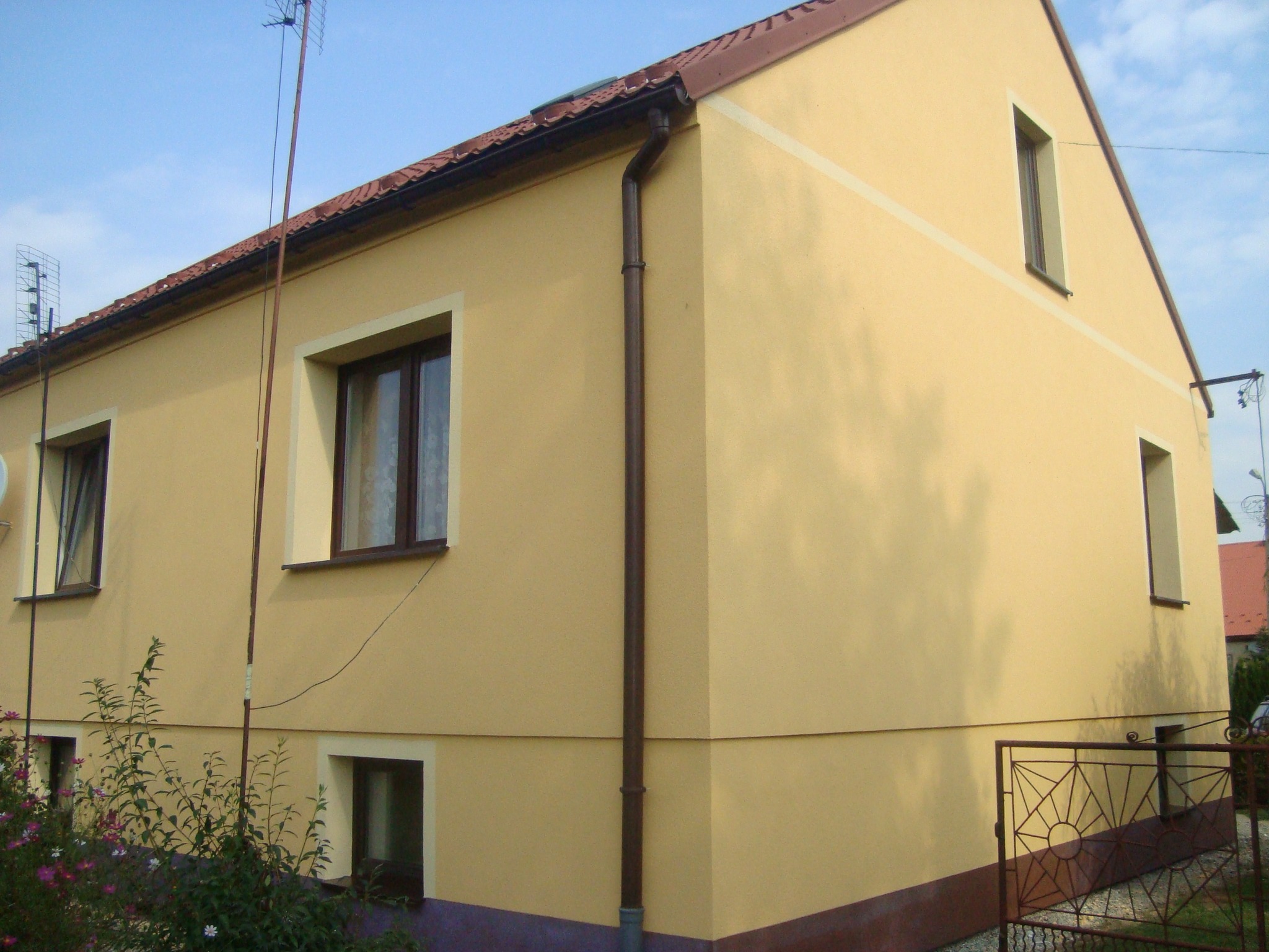 Żółta elewacja domu jednorodzinnego z brązowymi oknami i dachem, widok z zewnątrz na ścianę boczną i fragment frontu, z anteną na dachu i roślinnością u podstawy.