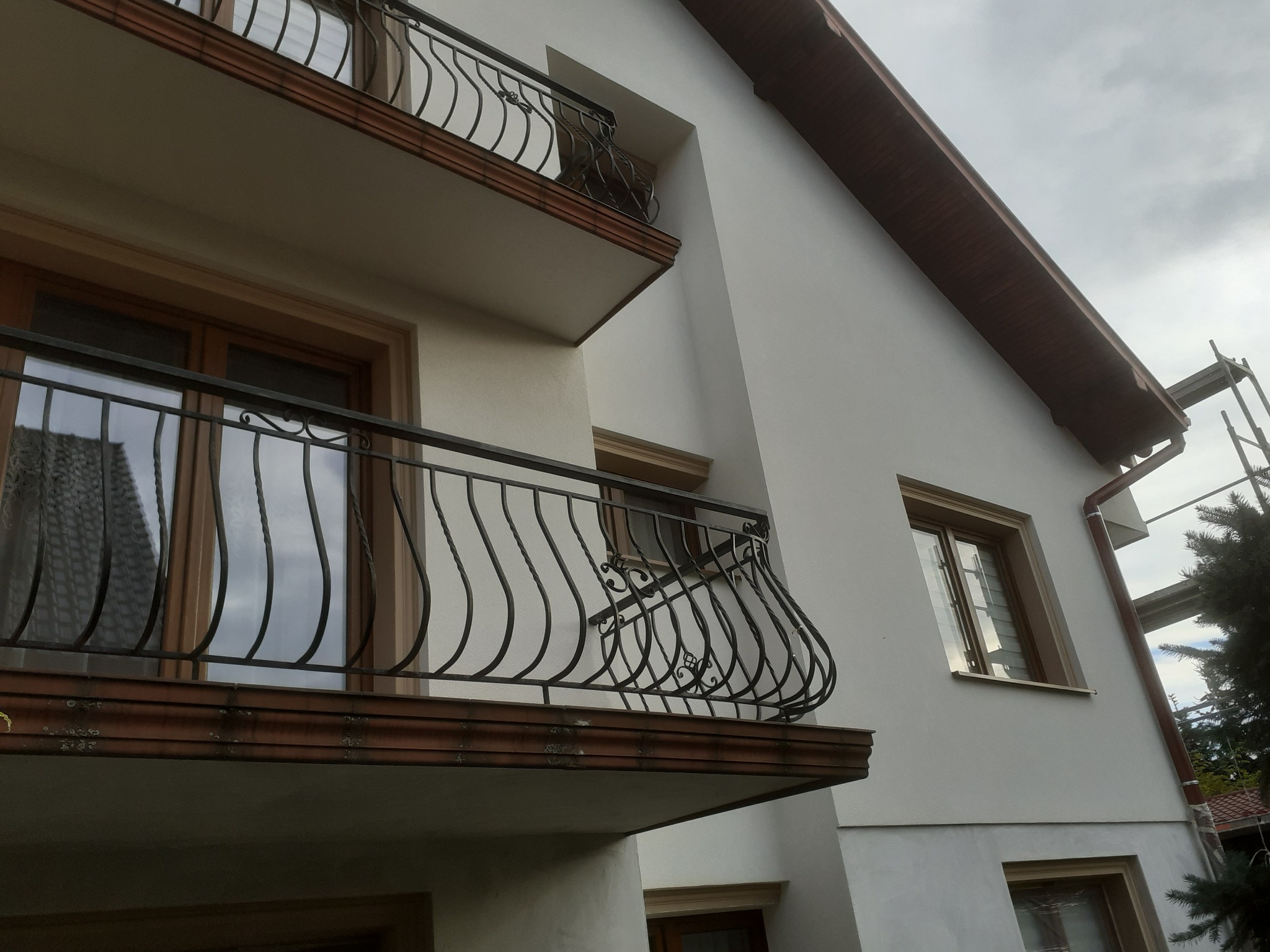Jasna elewacja domu z balkonami o zdobionych, kutych balustradach, widoczne okna z drewnianymi ramami i fragment rusztowania.