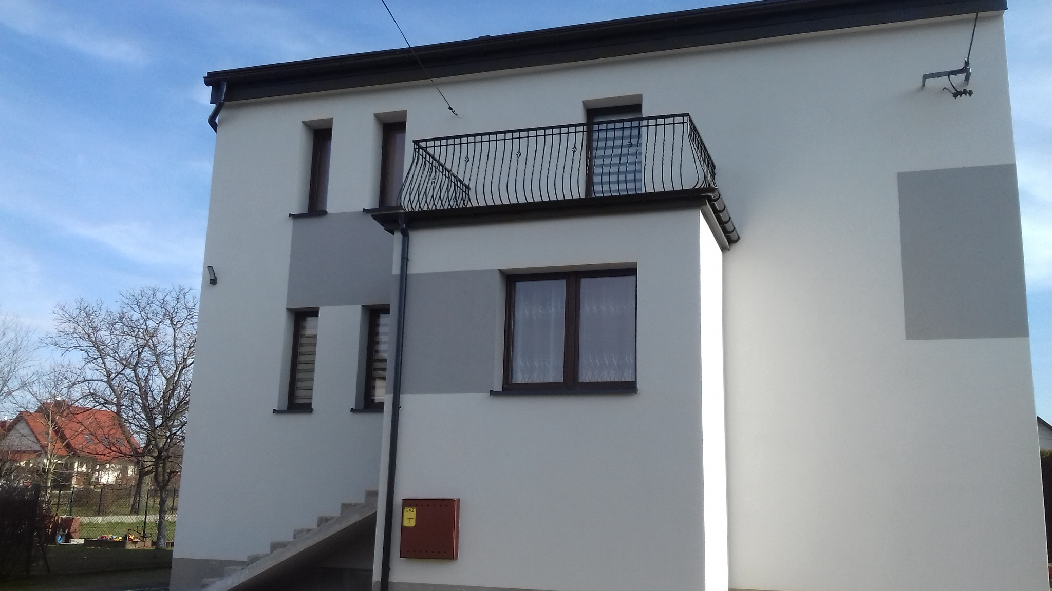Dwukondygnacyjny dom z balkonem z kutą balustradą, świeżo otynkowaną i pomalowaną elewacją w odcieniach bieli i szarości, z widoczną skrzynką gazową i drzewami w tle.