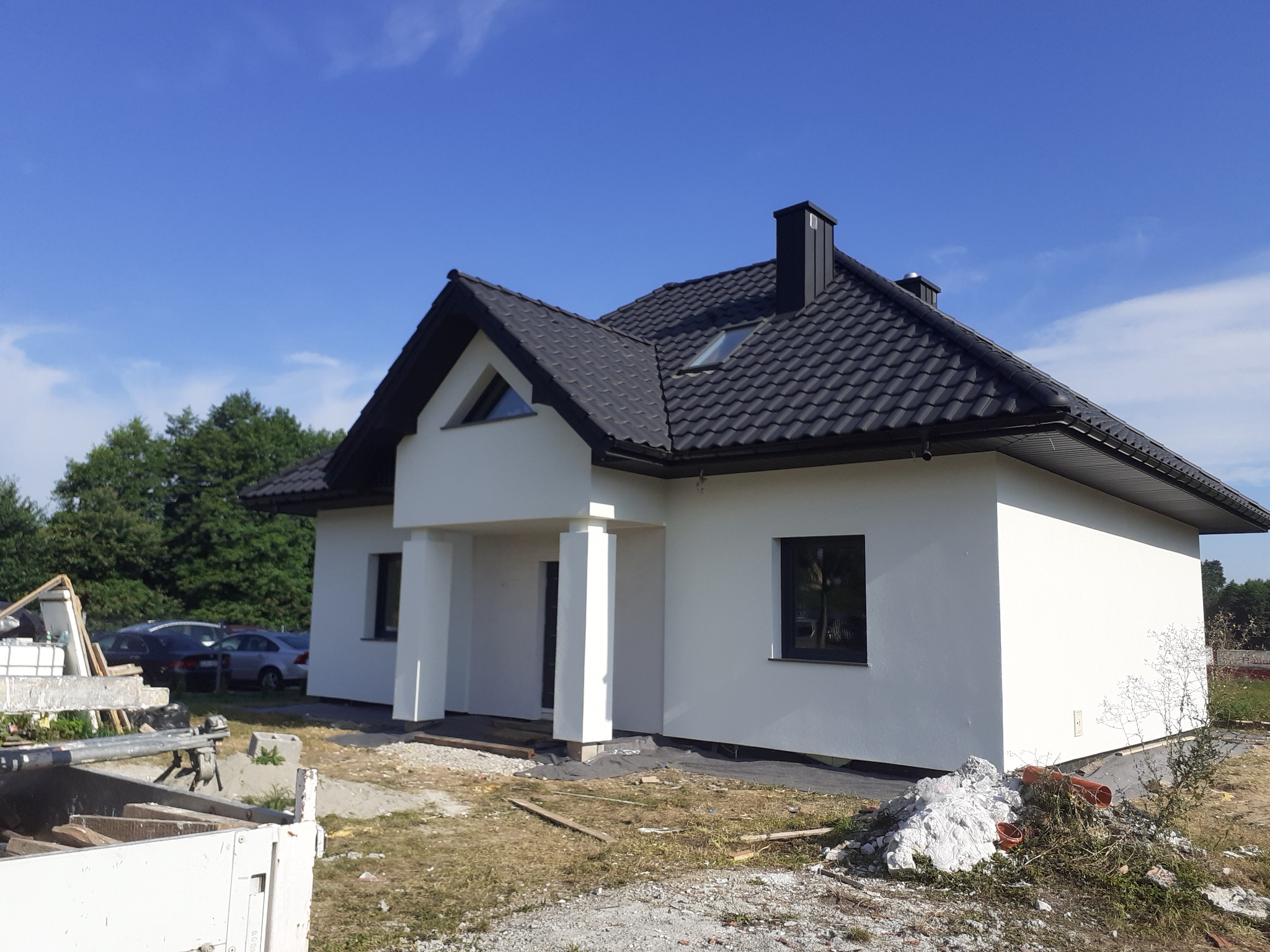 Nowoczesny dom jednorodzinny z białą elewacją, czarnym dachem i stolarką okienną, widoczny z zewnątrz, z niezagospodarowanym terenem wokół i stertą styropianu.
