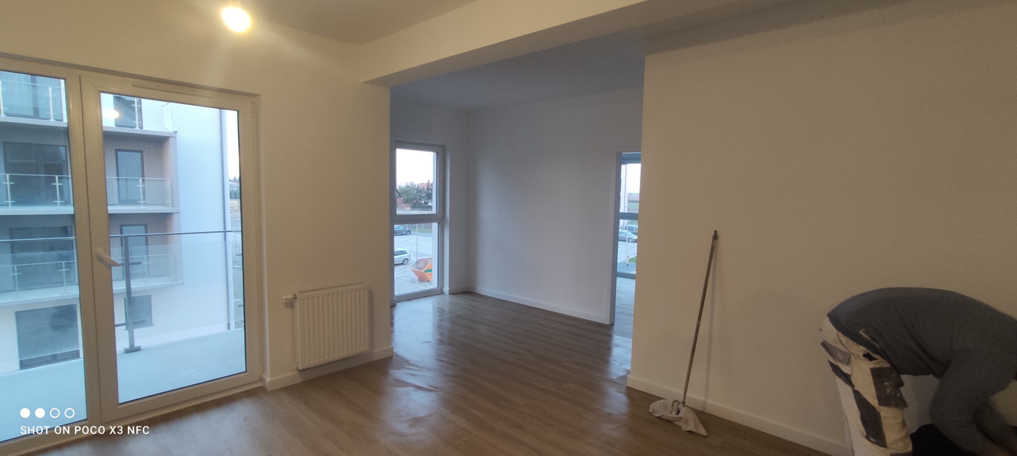 Wnętrze świeżo wykończonego mieszkania z widocznymi oknami balkonowymi, grzejnikiem, odnowioną podłogą i mopem opartym o ścianę, z fragmentem osoby w tle.