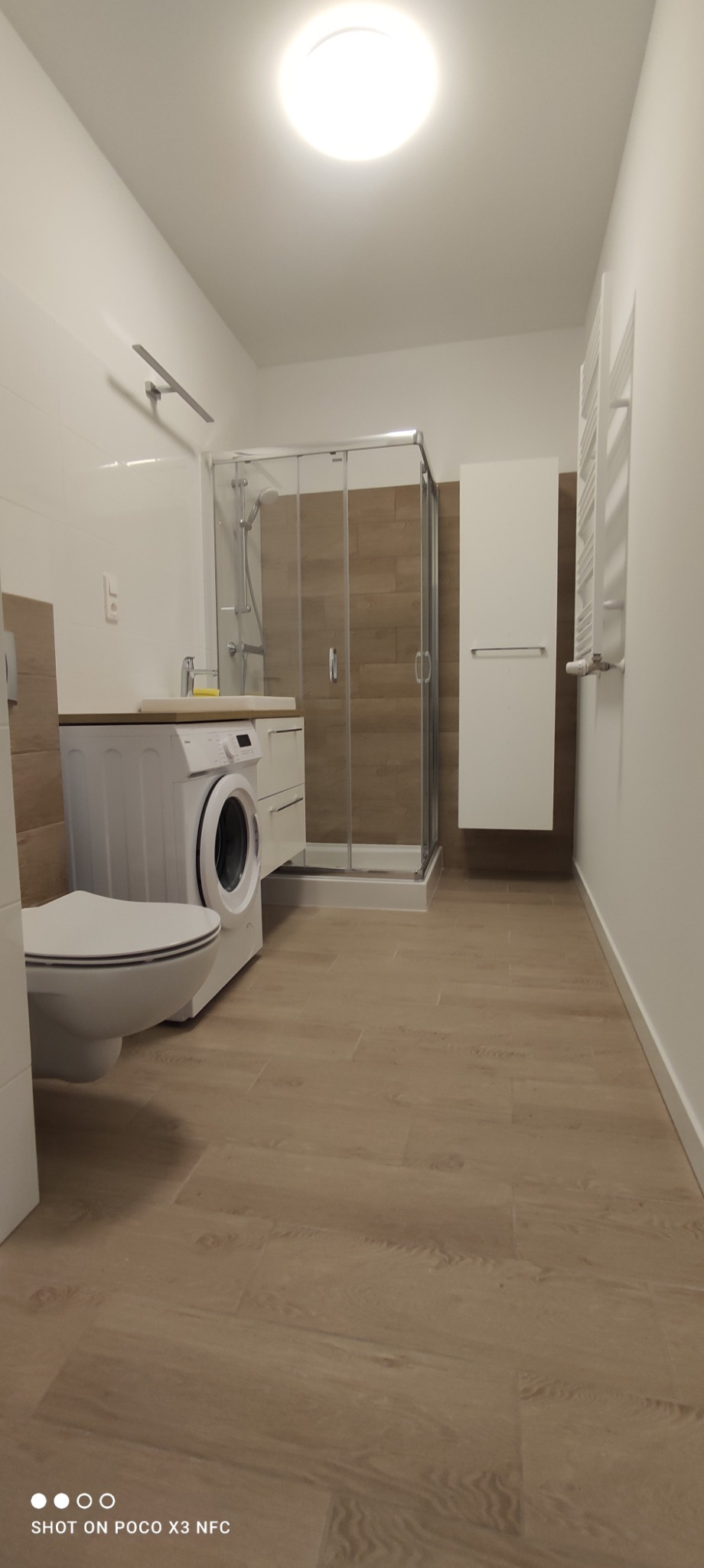 Nowoczesna łazienka po remoncie z kabiną prysznicową, pralką pod blatem umywalki i drewnopodobną podłogą z płytek.