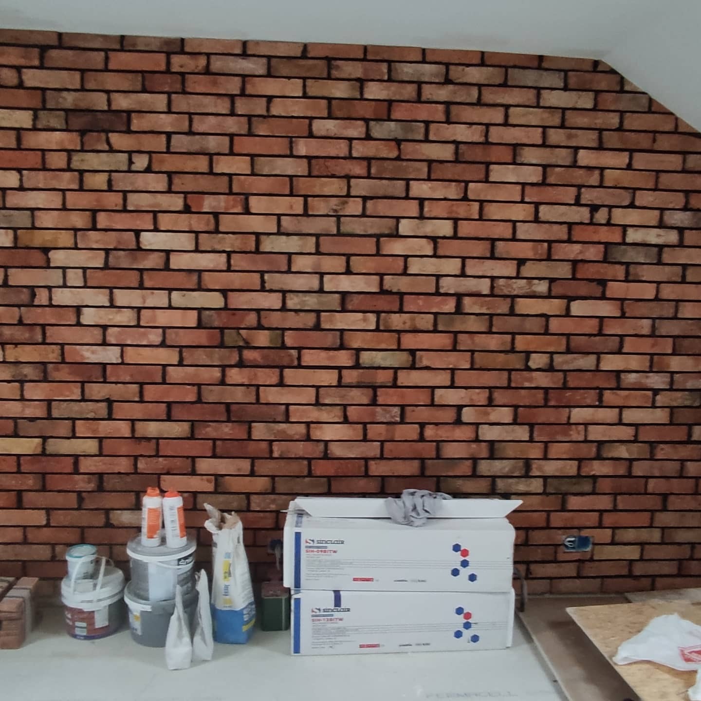 Ściana wykończona czerwoną cegłą klinkierową z widocznymi zapasami materiałów budowlanych, w tym worki z zaprawą i opakowania płyt izolacyjnych Sinclair.