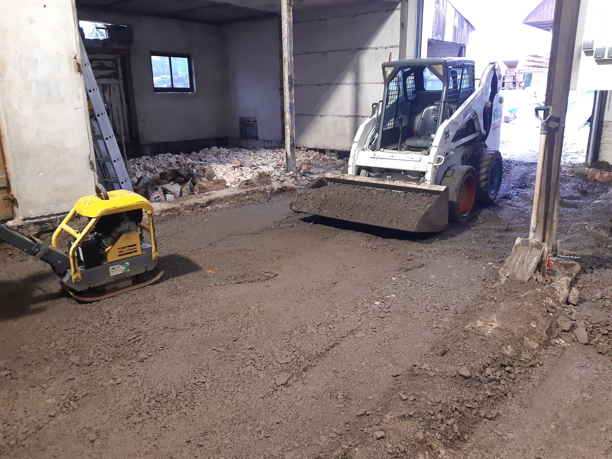 Przygotowanie podłoża pod posadzkę wewnątrz budynku: ubijarka wibracyjna i miniładowarka na gruncie z gruzem i ziemią.