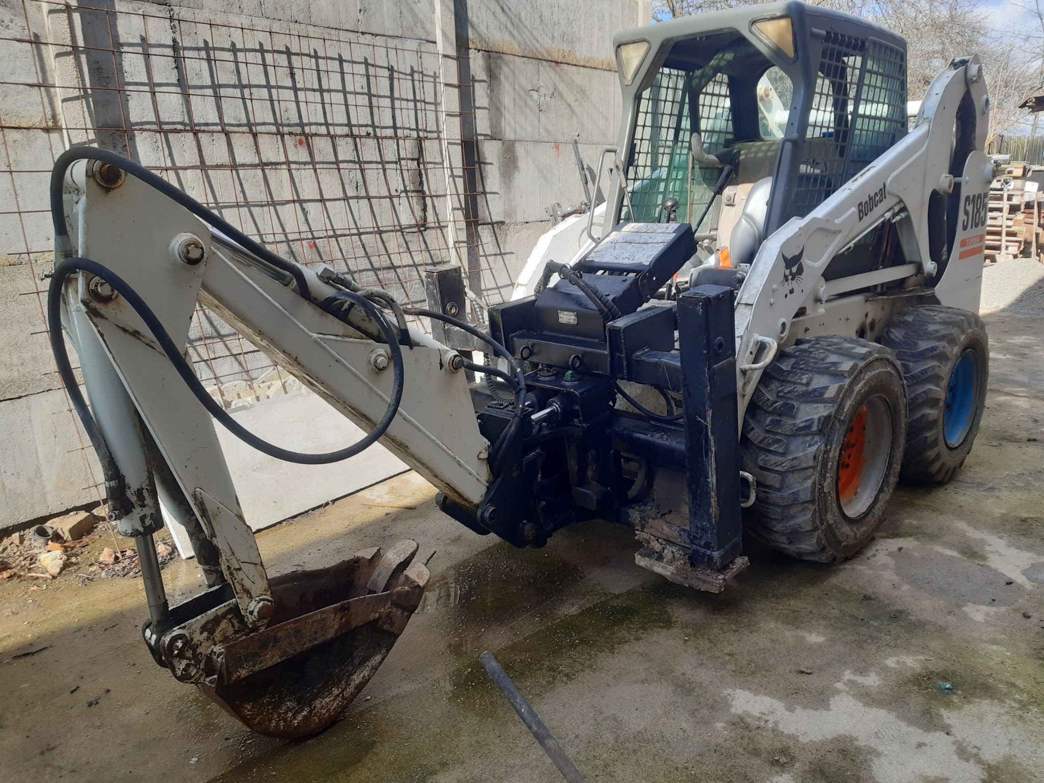 Biała, używana mini-ładowarka Bobcat S185 z łyżką koparkową, stojąca na betonowym podłożu z widoczną ścianą z siatką zbrojeniową w tle.