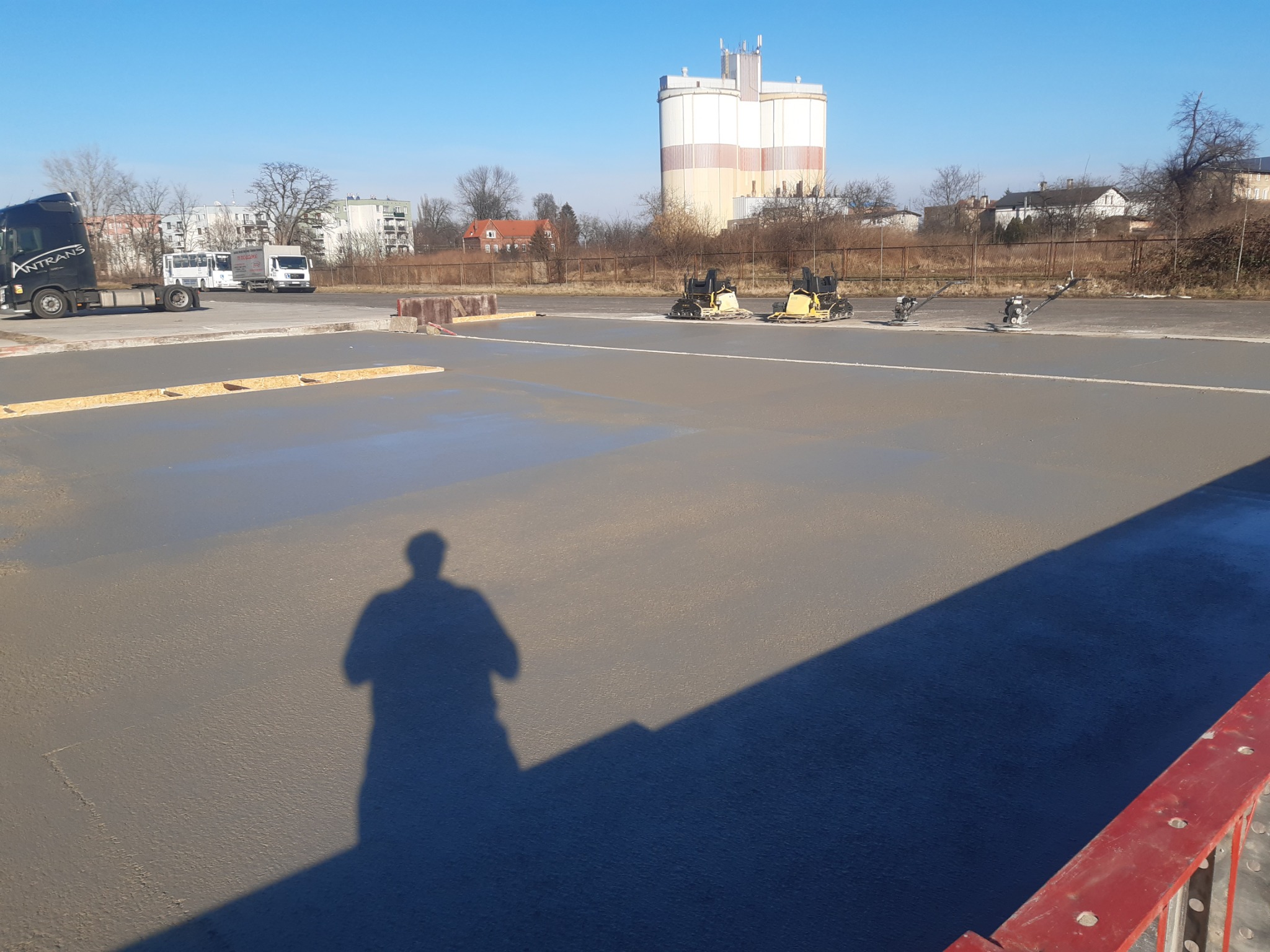 Świeżo wylana posadzka betonowa w trakcie wygładzania z użyciem zacieraczek mechanicznych, cień osoby na posadzce, w tle budynki i silos.