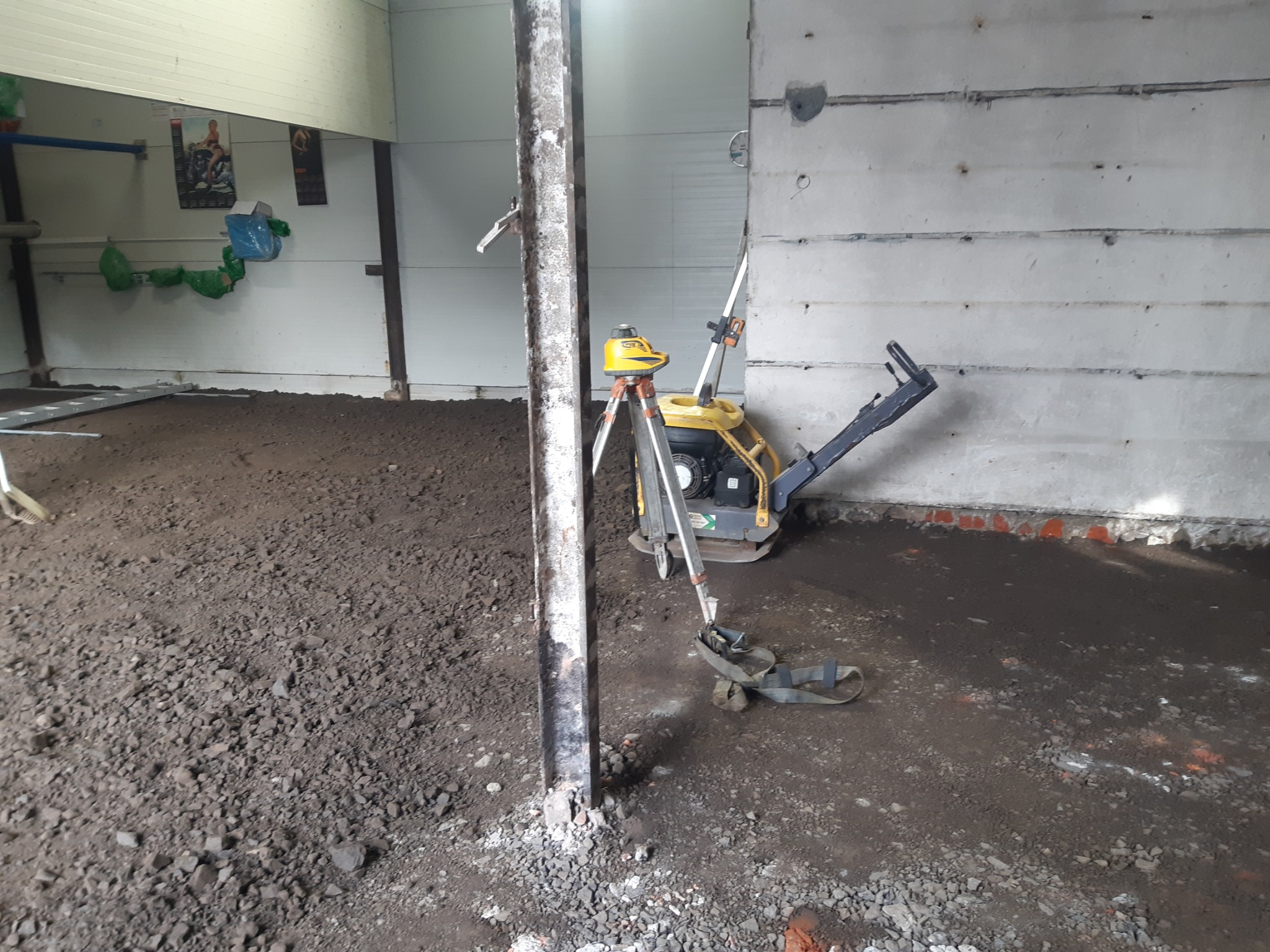 Przygotowanie podłoża pod posadzkę w hali: ubijarka wibracyjna i niwelator laserowy ustawione na nierównym, gruzowym podłożu, widoczna stalowa kolumna konstrukcyjna.