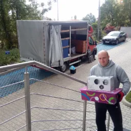 Mężczyzna w szarej bluzie i czerwonych rękawicach trzyma pudełka przed otwartą ciężarówką załadowaną meblami podczas przeprowadzki.