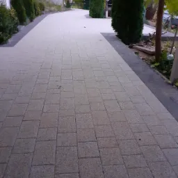 Szeroka, jasnoszara nawierzchnia z kostki brukowej prowadząca wzdłuż posesji z iglakami i budynkiem w tle, wykończona ciemniejszym obrzeżem.