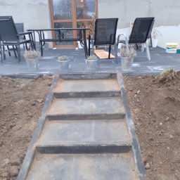 ROBUD - Schody zewnętrzne z ciemnych płyt prowadzące na taras wyłożony kostką, z widocznymi doniczkami z lawendą i meblami ogrodowymi na tle domu z dużymi oknami.