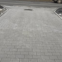 ROBUD - Nowo ułożona szara kostka brukowa przed posesją, widoczna krawędź jezdni i fragment samochodu. Kostka ułożona w regularny wzór, z widocznymi spoinami. Część kostki jeszcze nieułożona.