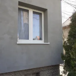 Świeżo otynkowana szara elewacja budynku z białym oknem, widoczne nierówności faktury tynku, dolna część ściany wykończona kamieniem elewacyjnym, obok iglak.