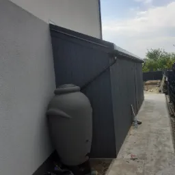 Szara beczka na deszczówkę z podłączonym spustem rynnowym, stojąca przy ścianie budynku obok ciemnoszarej drewnianej wiaty.