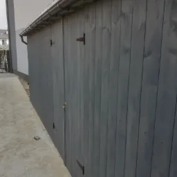 Szare drewniane ogrodzenie z metalowymi zawiasami, widok z boku, z rynną wzdłuż górnej krawędzi, obok wyłożona płytami ścieżka.