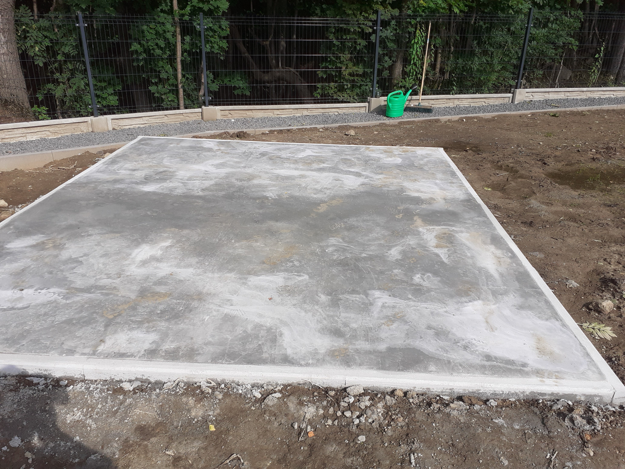 Wylana płyta betonowa pod budowę, otoczona obrzeżem, widoczny fragment ogrodzenia panelowego i zielony trawnik z drzewami w tle.