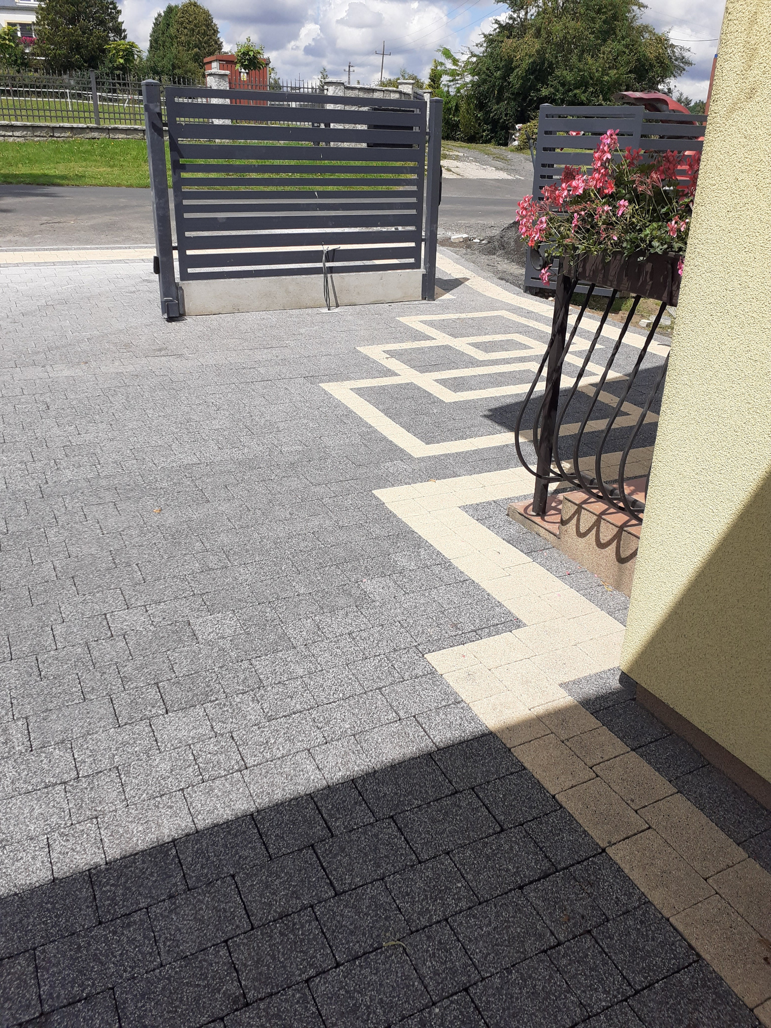 Szara kostka brukowa ułożona we wzór z jasnymi akcentami, widoczna brama wjazdowa z poziomymi szczeblami i donica z różowymi kwiatami przy ścianie budynku.