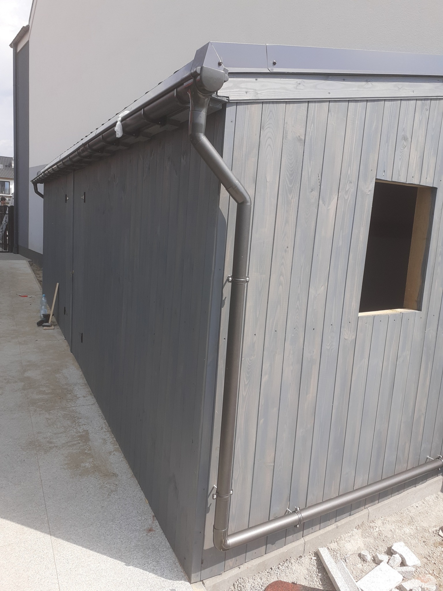 Szary, drewniany budynek gospodarczy z rynną, widoczne okno bez szyby i betonowa ścieżka.