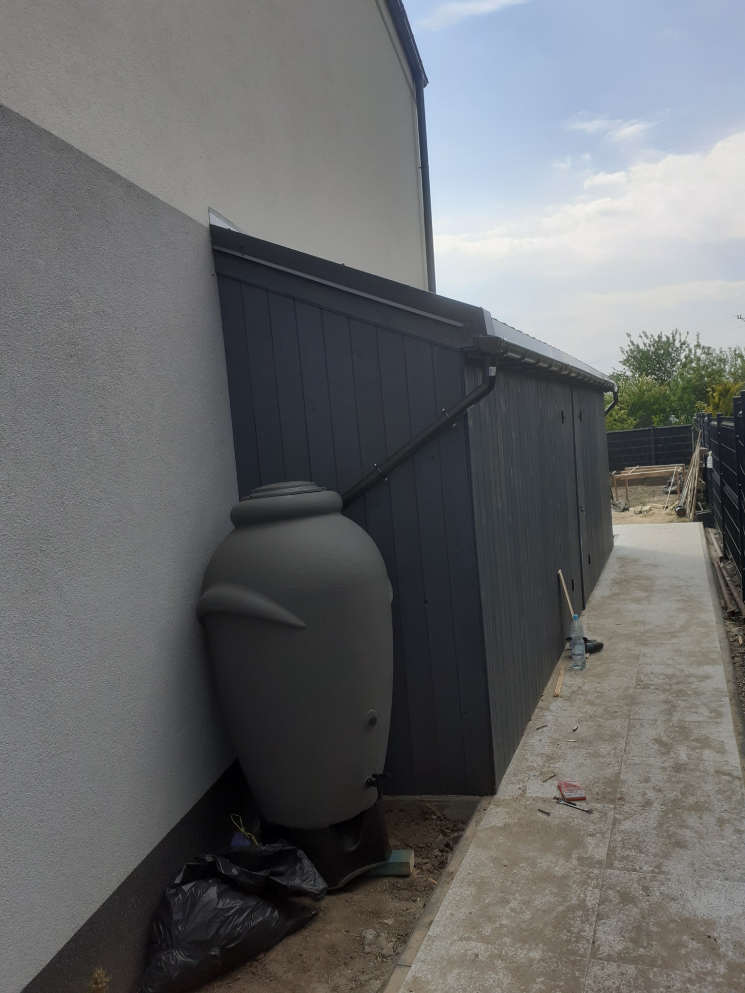 Szara beczka na deszczówkę z podłączonym spustem rynnowym, stojąca przy ścianie budynku obok ciemnoszarej drewnianej wiaty.