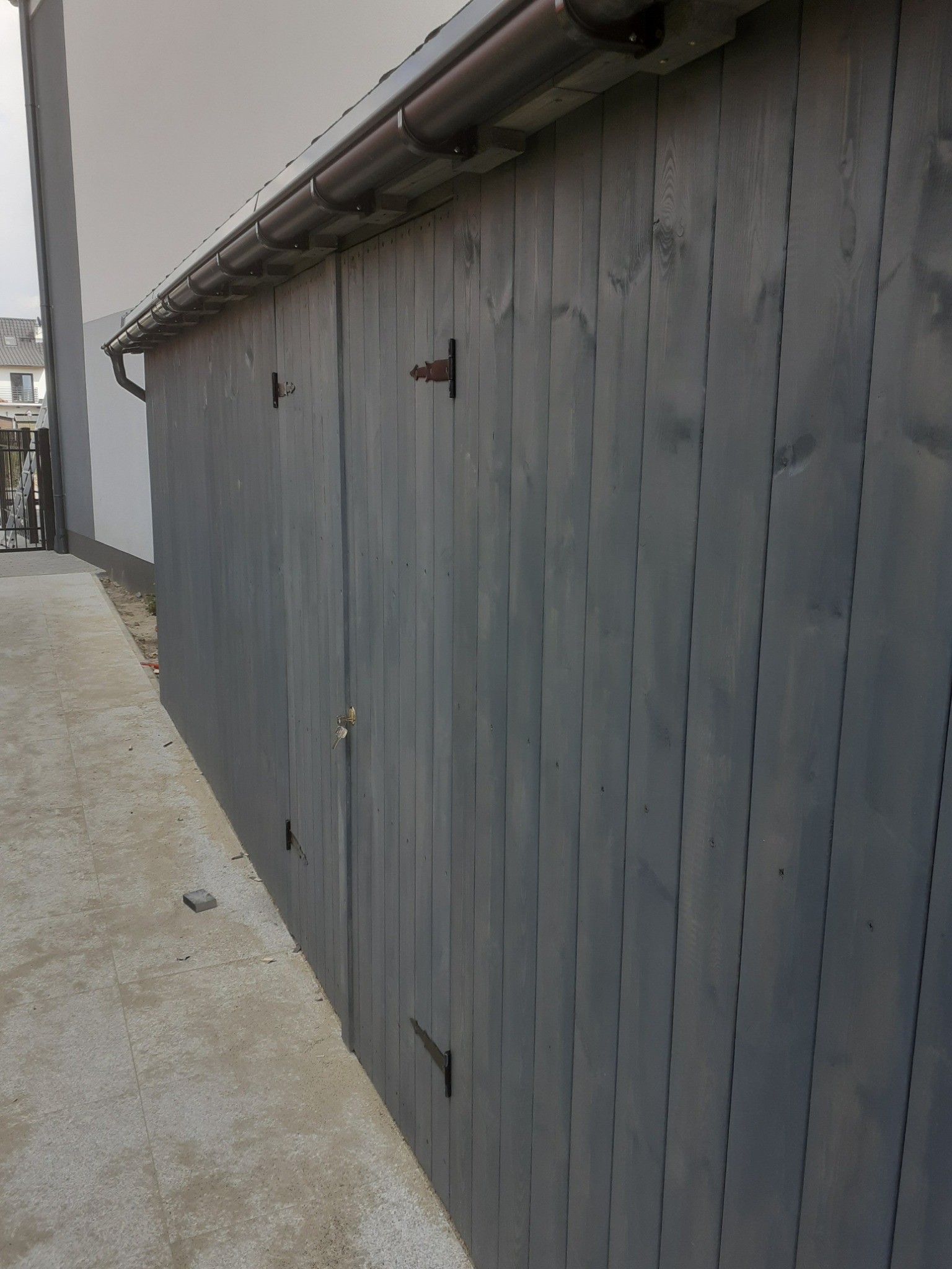 Szare drewniane ogrodzenie z metalowymi zawiasami, widok z boku, z rynną wzdłuż górnej krawędzi, obok wyłożona płytami ścieżka.