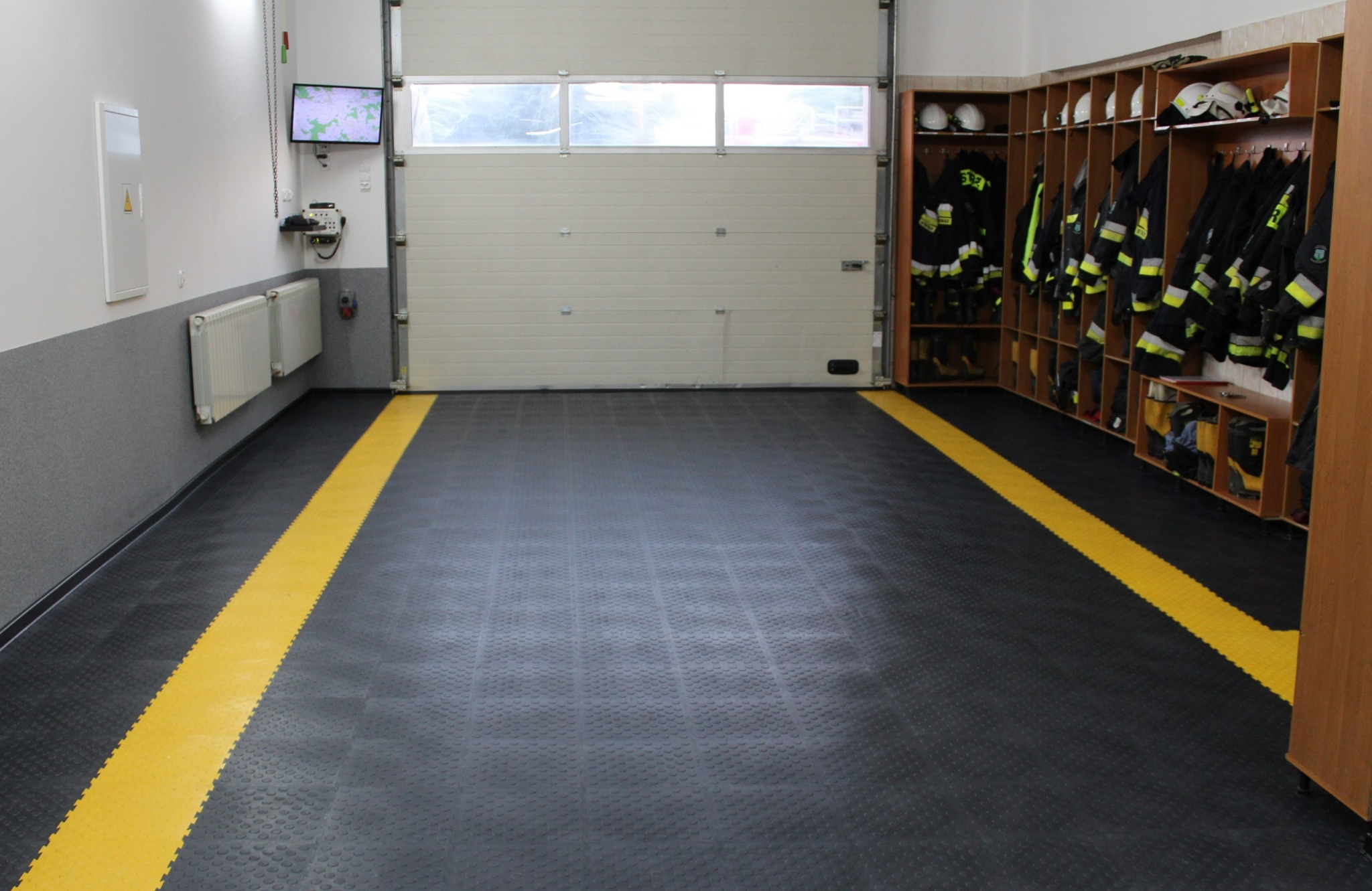 Garaż strażacki z czarną podłogą z żółtymi pasami, białą bramą garażową i szafą na ubrania ochronne