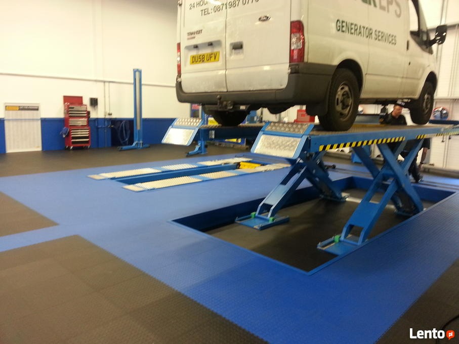 Warsztat samochodowy z podnośnikiem i busem, widoczny pracownik pod pojazdem, niebiesko-czarna podłoga z gumowych płyt.
