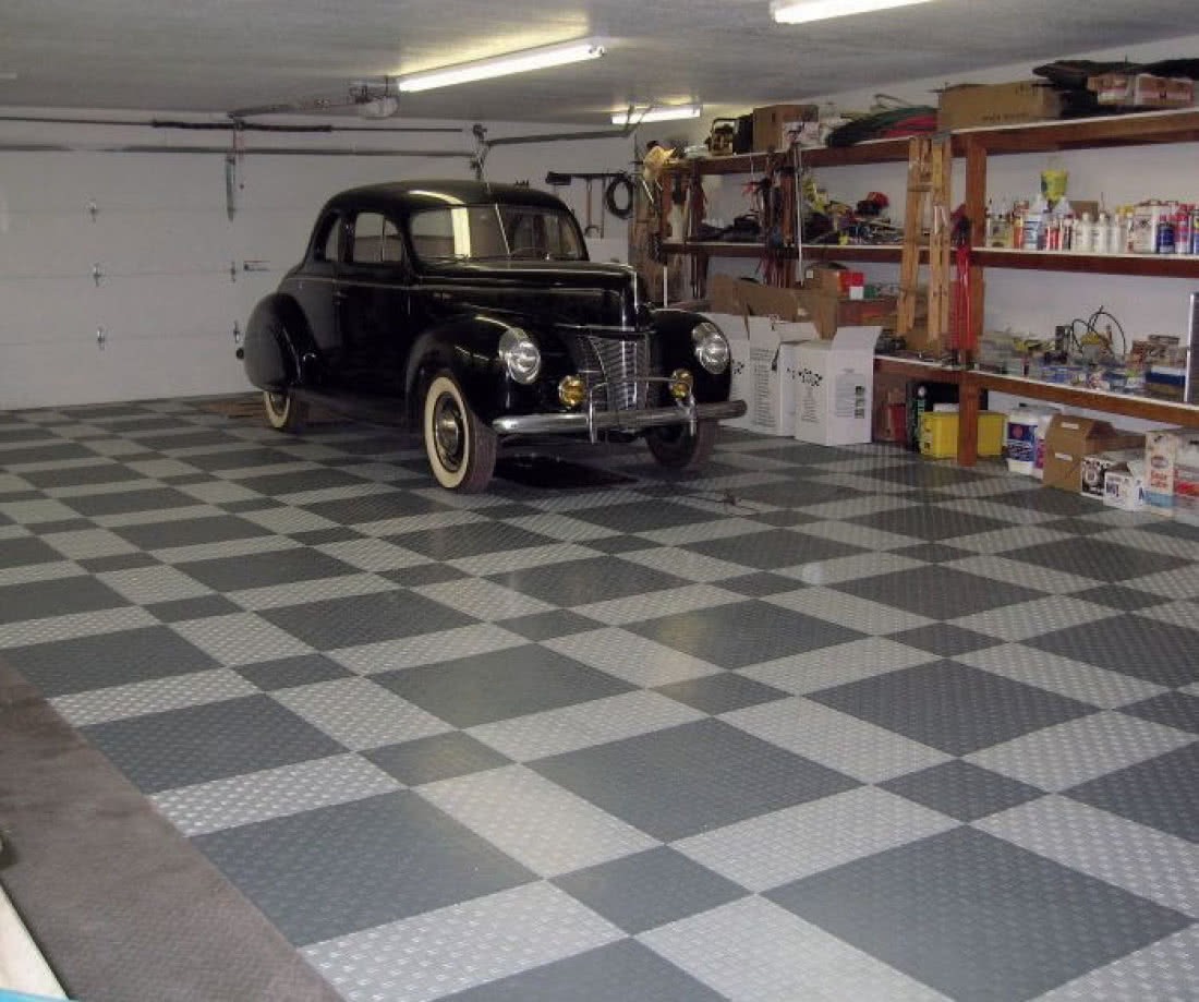 Szara podłoga w szachownicę w garażu z czarnym klasycznym samochodem i półkami z narzędziami w tle.