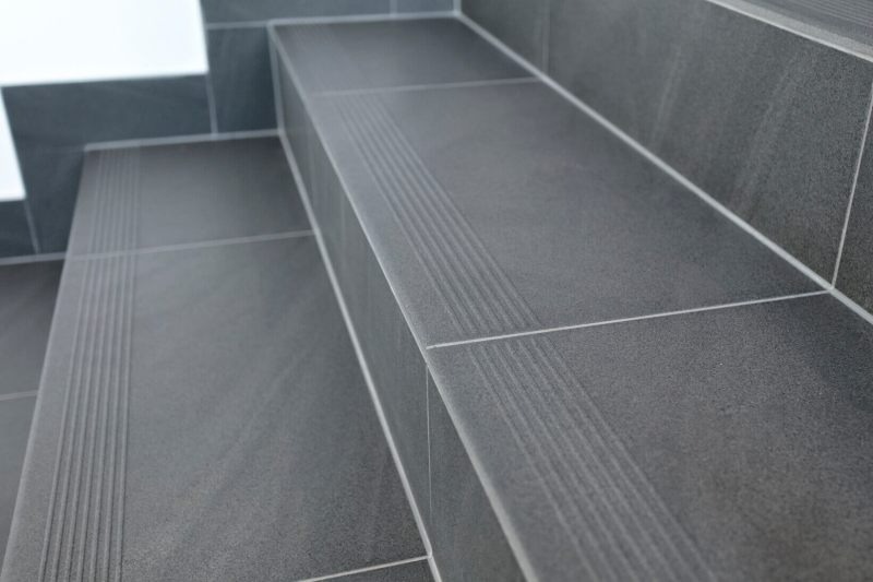 Schody wyłożone ciemnoszarą, matową glazurą z subtelnymi, prążkowanymi detalami antypoślizgowymi na krawędziach stopni, wykończone jasną fugą.