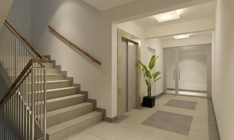 Jasne, nowocześnie wykończone wnętrze klatki schodowej z windą, schodami z metalową balustradą i drewnianym poręczą, rośliną doniczkową oraz dwuskrzydłowymi drzwiami.