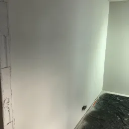 Świeżo wykończona, gładka ściana w rogu pokoju, zabezpieczona folią podłoga, widoczne puszki elektryczne.