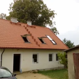 Dom jednorodzinny z nową, czerwoną dachówką falistą, trzema oknami dachowymi i dwoma kominami, widoczny z poziomu gruntu na tle drzew i pochmurnego nieba.