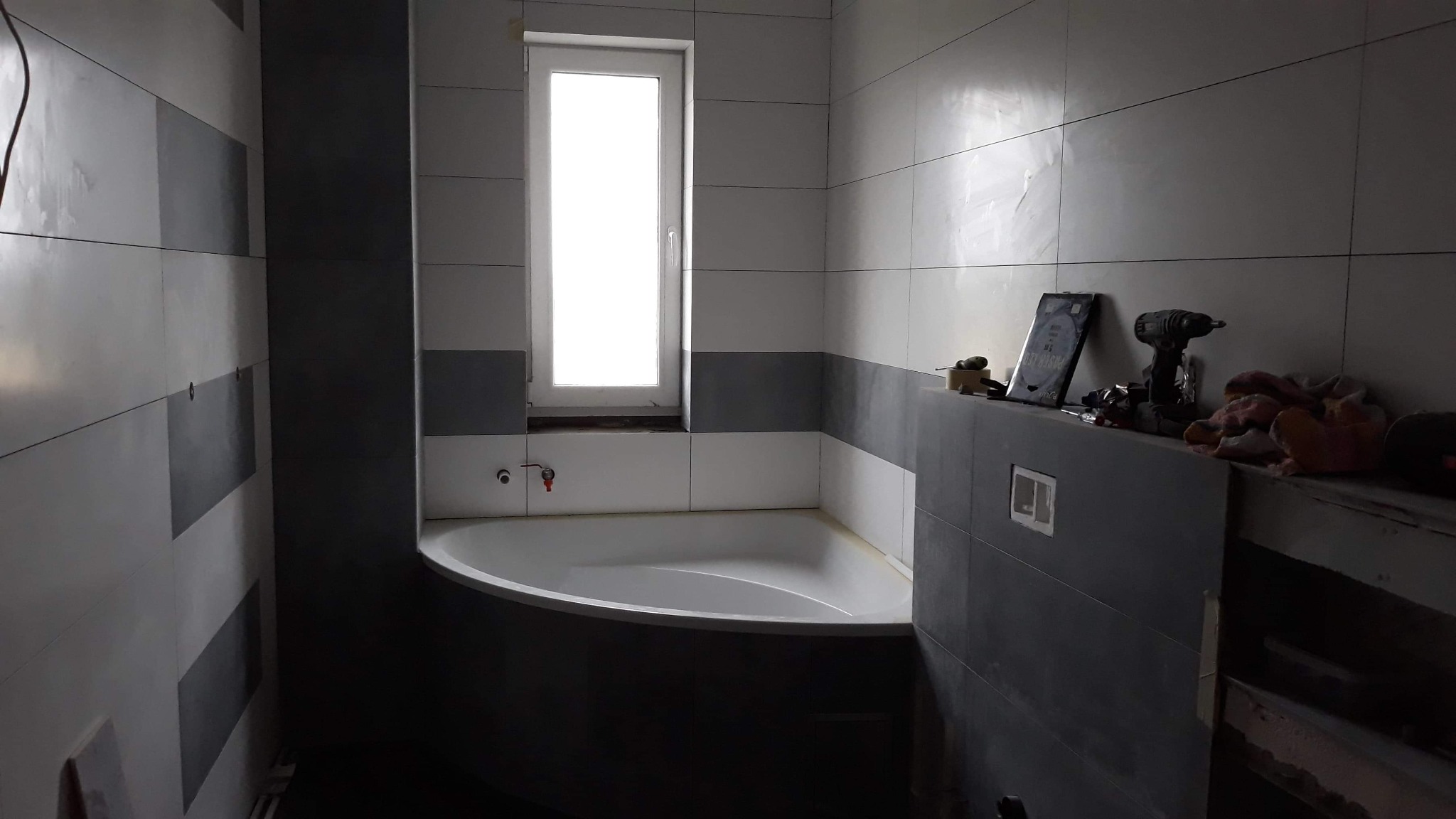Wnętrze łazienki w trakcie prac wykończeniowych; widoczna wanna narożna, okno, ściany z szarymi i białymi płytkami oraz narzędzia budowlane na półce.