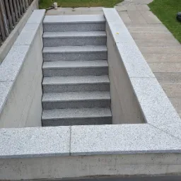 Zewnętrzne schody z szarego granitu prowadzące do piwnicy, otoczone betonową ramą wyłożoną granitem, widoczne ogrodzenie drewniane i trawnik