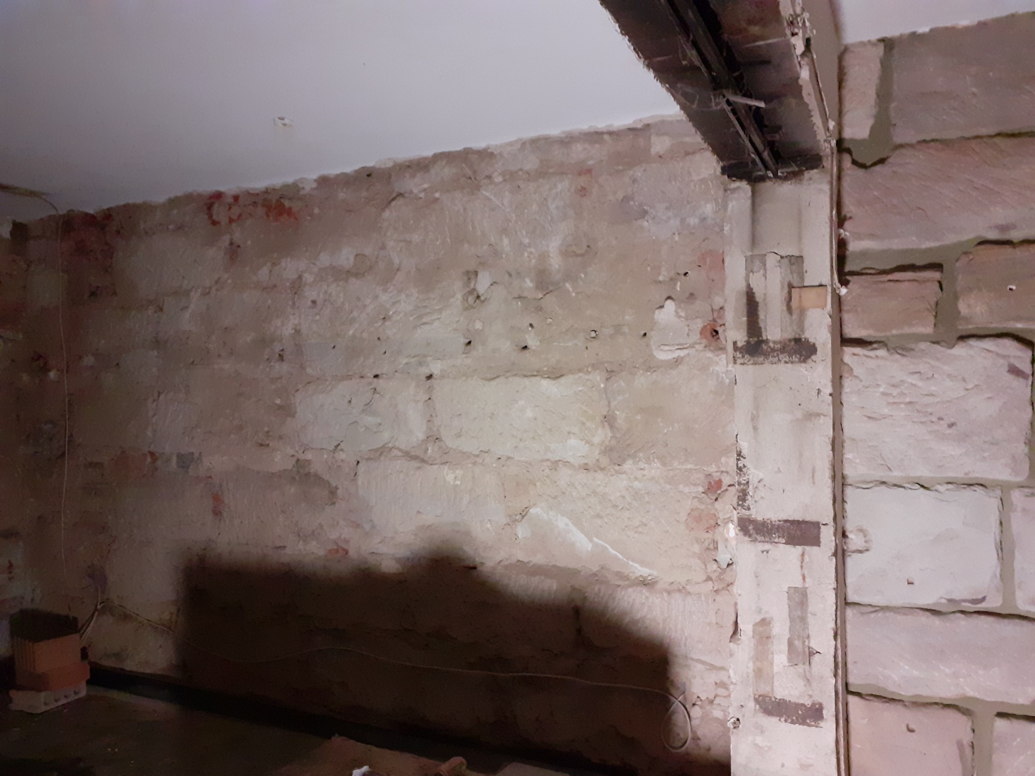 Surowa ściana z odspojonym tynkiem, fragment muru z cegły i instalacja pod sufitem w trakcie prac remontowych.
