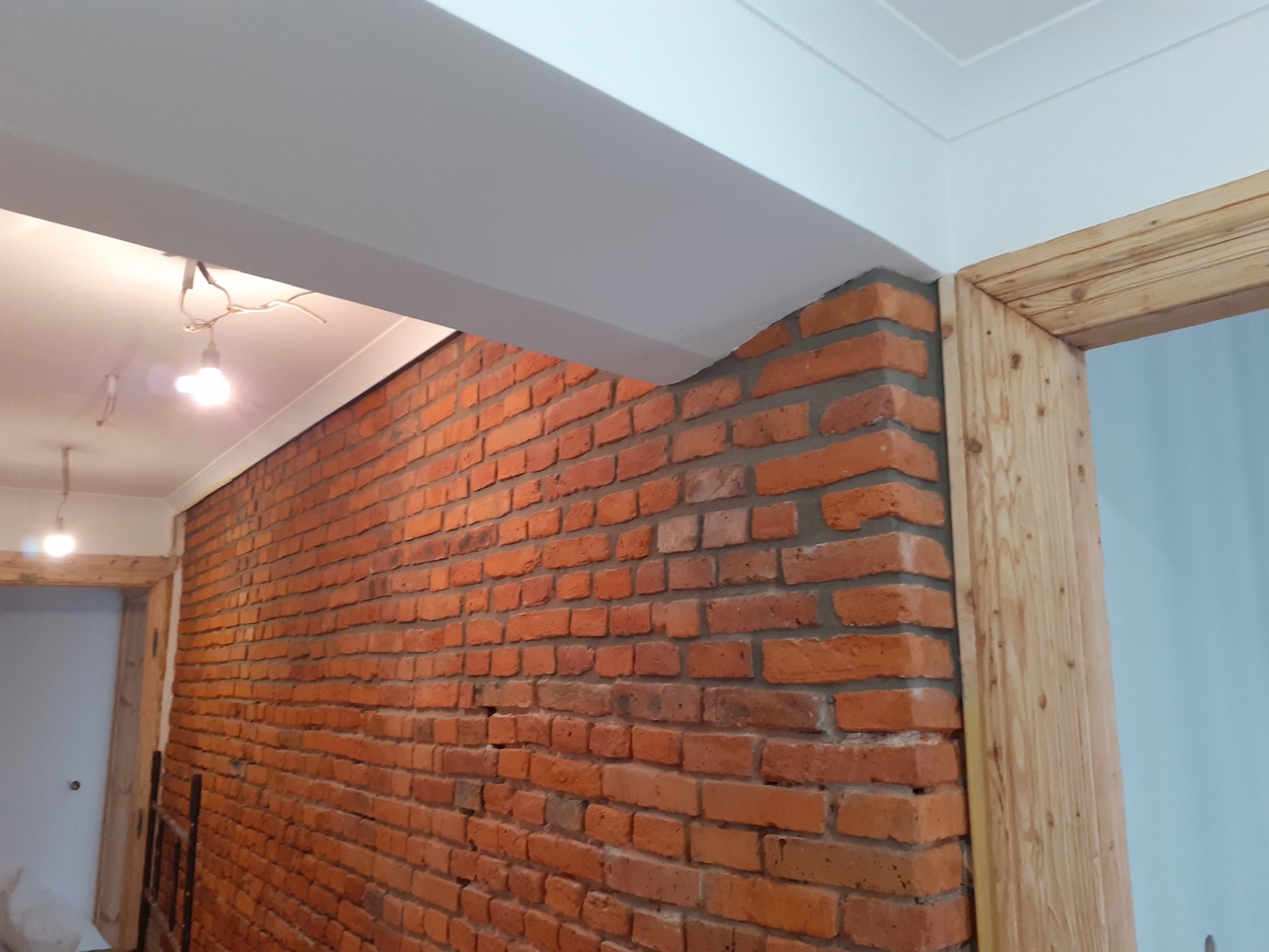 Czerwona ściana z cegły połączona szarą zaprawą, obok drewniana ościeżnica drzwi, biały sufit z wystającymi kablami żarówki, widok z dołu.