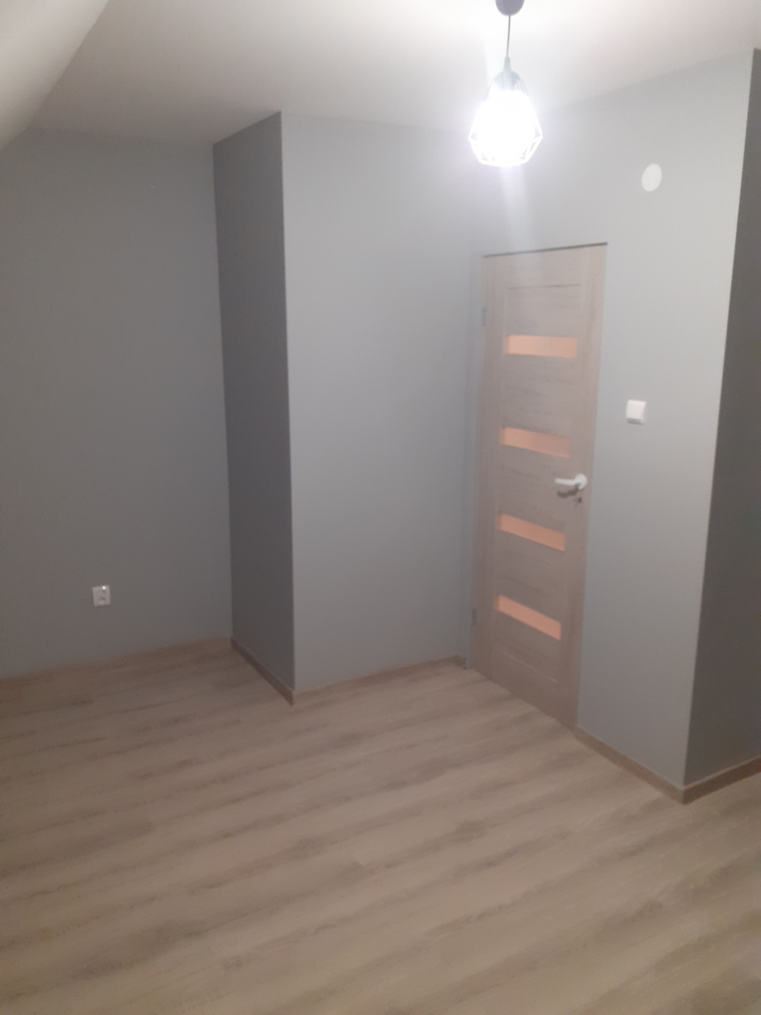 Pokój po remoncie: szare ściany, drewniana podłoga, jasne drzwi z przeszkleniami, widoczny włącznik światła i gniazdko elektryczne, oświetlenie sufitowe.