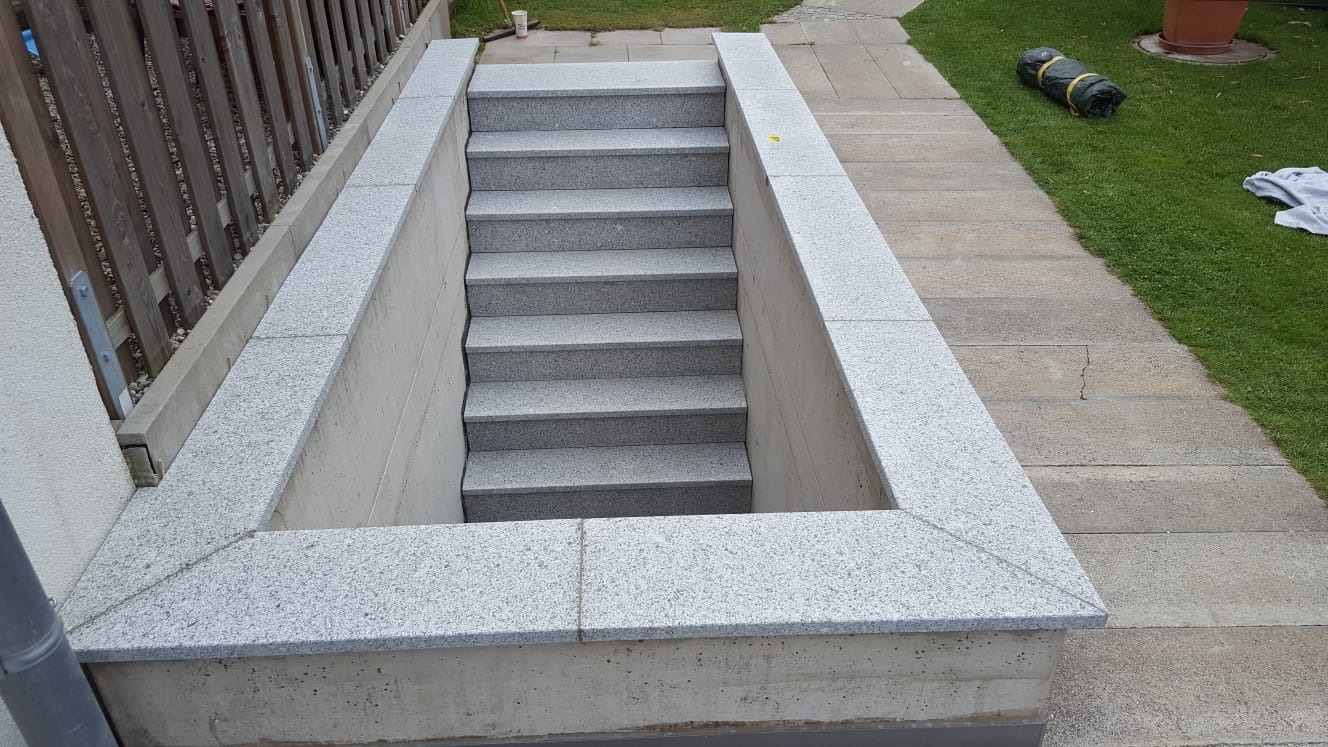 Zewnętrzne schody z szarego granitu prowadzące do piwnicy, otoczone betonową ramą wyłożoną granitem, widoczne ogrodzenie drewniane i trawnik