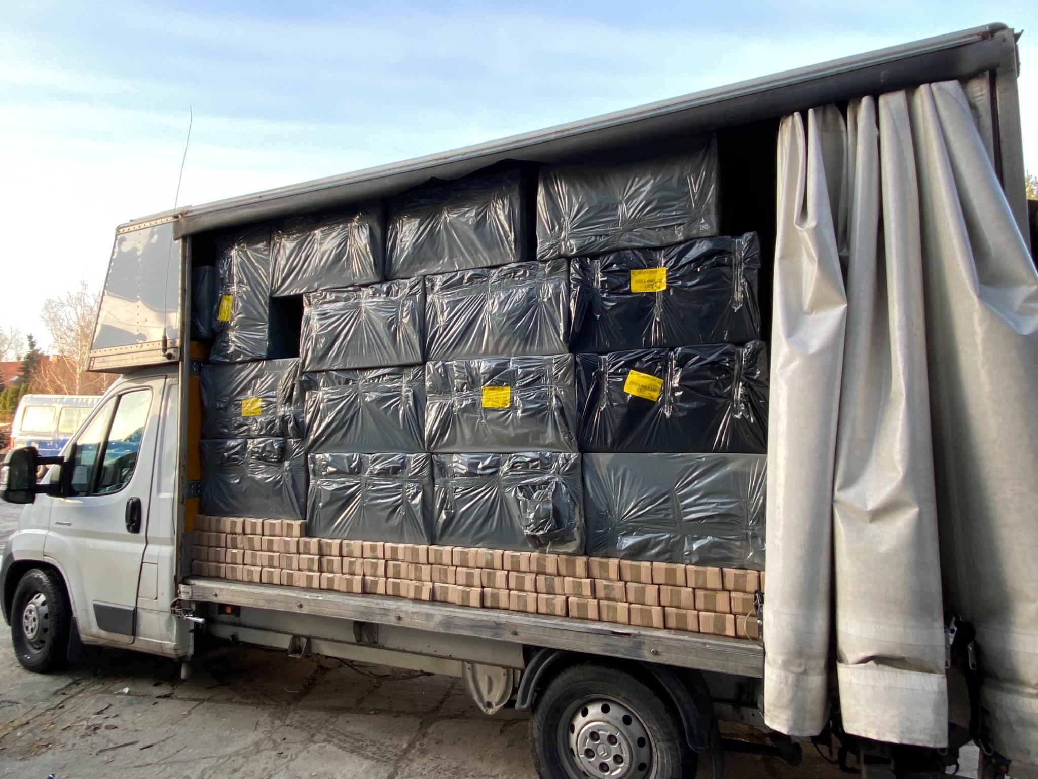 Bus transportuje palety cegieł i zapakowane w folię towary z żółtymi etykietami w przestrzeni ładunkowej z częściowo odsłoniętą plandeką.