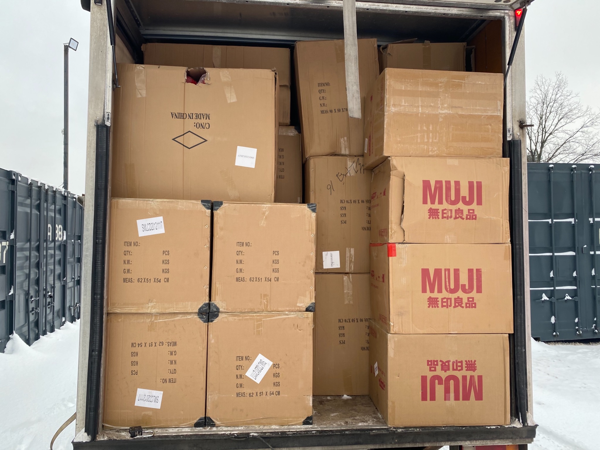Wnętrze furgonetki wypełnionej kartonami różnych rozmiarów, niektóre z logo MUJI, widoczne etykiety z informacjami o produktach, tło stanowi zimowy krajobraz z kontenerami.