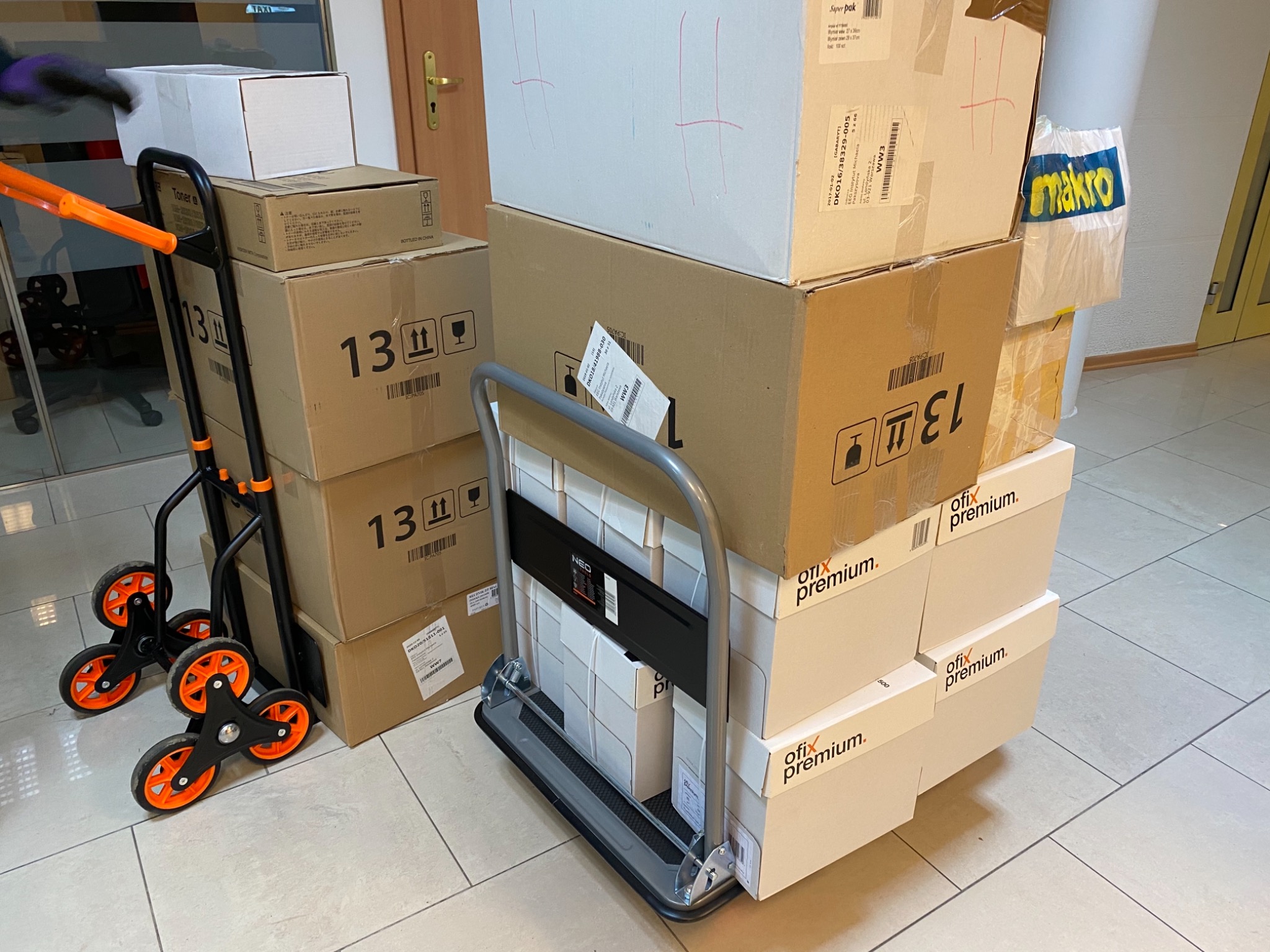 Dwa wózki transportowe załadowane kartonami różnej wielkości, gotowe do przewiezienia w biurze.