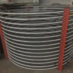 Spiralna rura ze stali nierdzewnej, podparta czerwonymi profilami, widoczna w warsztacie.