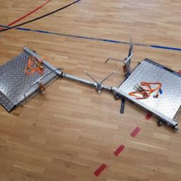 Niko-force sp. z o.o. - Dwa metalowe podesty z kratownicą, połączone rurą, z pomarańczowymi pasami mocującymi, ustawione na drewnianej podłodze hali sportowej z liniami boiska.