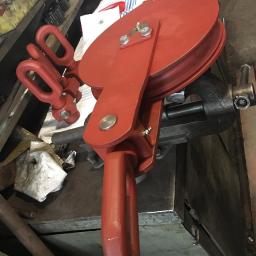 Niko-force sp. z o.o. - Czerwony bloczek linowy z trzema uchwytami, zamocowany w imadle na warsztatowym stole. Widoczne ślady użytkowania i warsztatowy nieład.