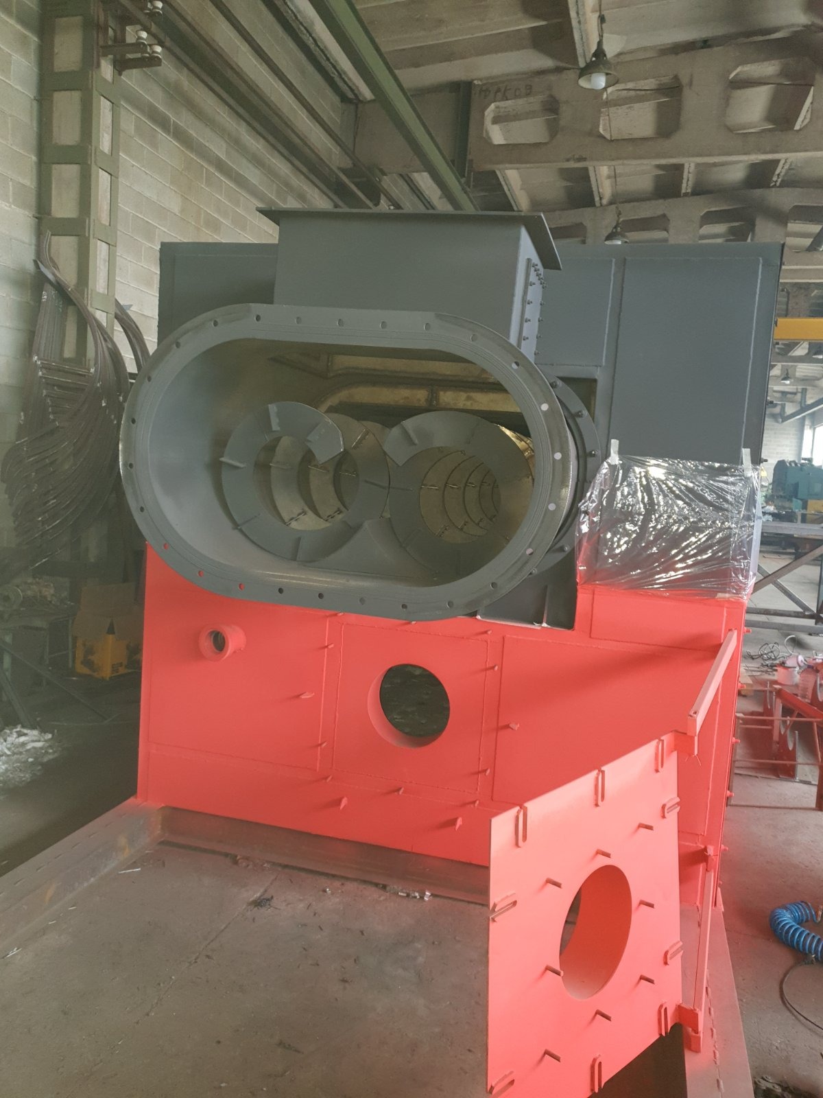 Częściowo złożona, pomalowana na szaro i pomarańczowo konstrukcja metalowa o dużych rozmiarach, z widocznymi elementami obrotowymi wewnątrz owalnego otworu, stojąca w hali produkcyjnej.